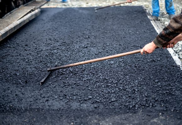 Worker leveling fresh asphalt on a road construction site, industrial buildings